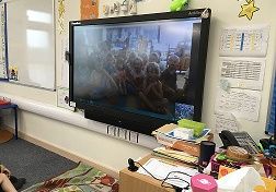 Na zdjęciu widać klasę dzieci z Wielkiej Brytanii po środku której jest duży ekran na którym widać dzieci.